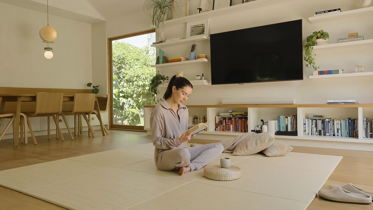A woman in loungewear sits cross-legged on a light modular tatami mat in a modern living room, reading a book.